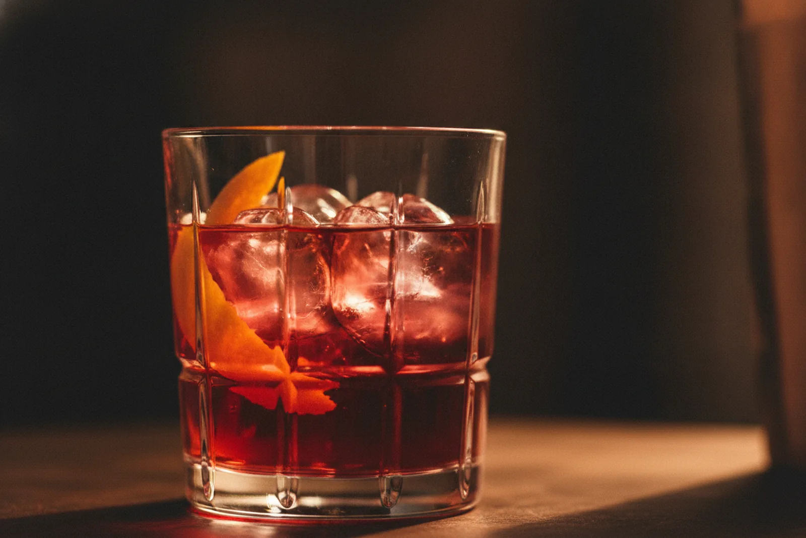 Ein Glas rubinroter Bitter-Aperitif mit Orangenzeste und Eis vor dunklem Hintergrund