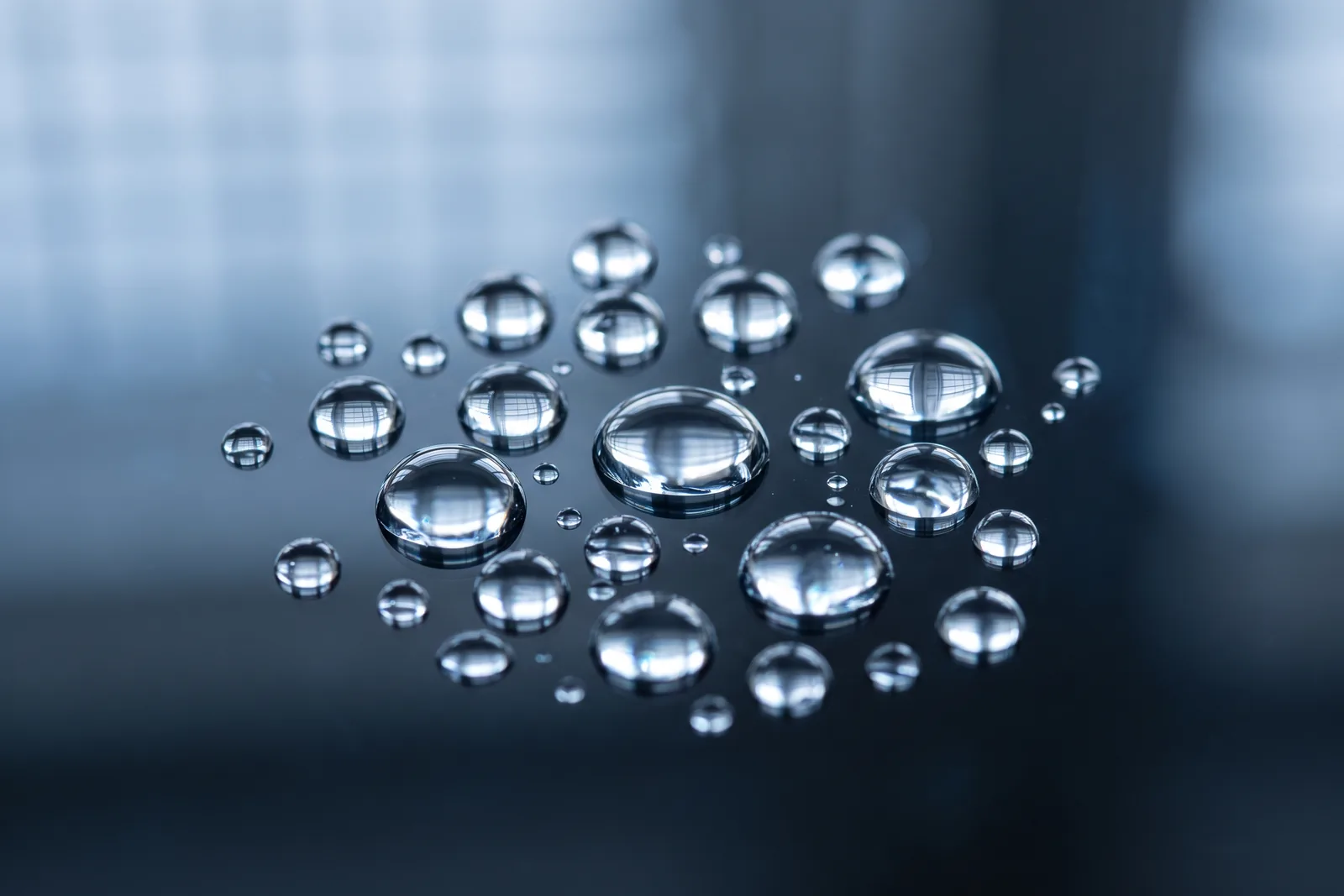 Klare Tropfen auf einer Glasoberfläche in kühlem Licht als Bild für die Präzision synthetischer Duftbausteine
